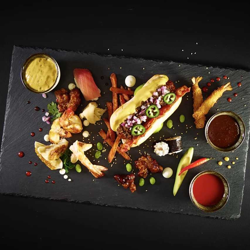 Hot Dog mit Yankee Hot Dog Sauce, Fire Cracker Sauce, Bacon, Zwiebeln, Jalapeños, Avocado, Edamame, veganer Mayo, Süßkartoffelpommes und Knuspergarnelen